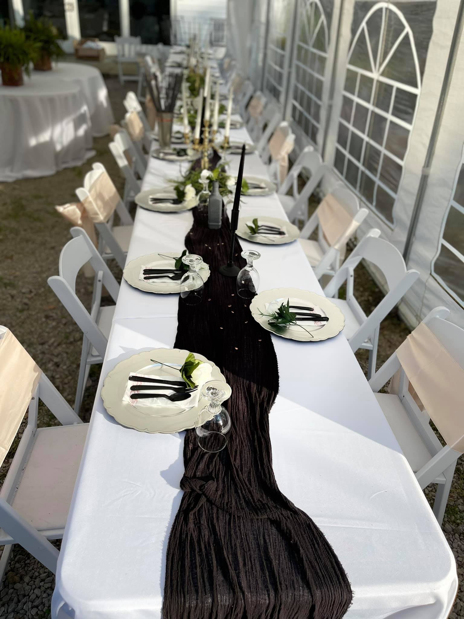 Photo of lon tale at reception with black table runner on white tablecloth candle centerpieces along the entire banquet and place settings with roses. Every other chair is wrapped with peach backing cloth