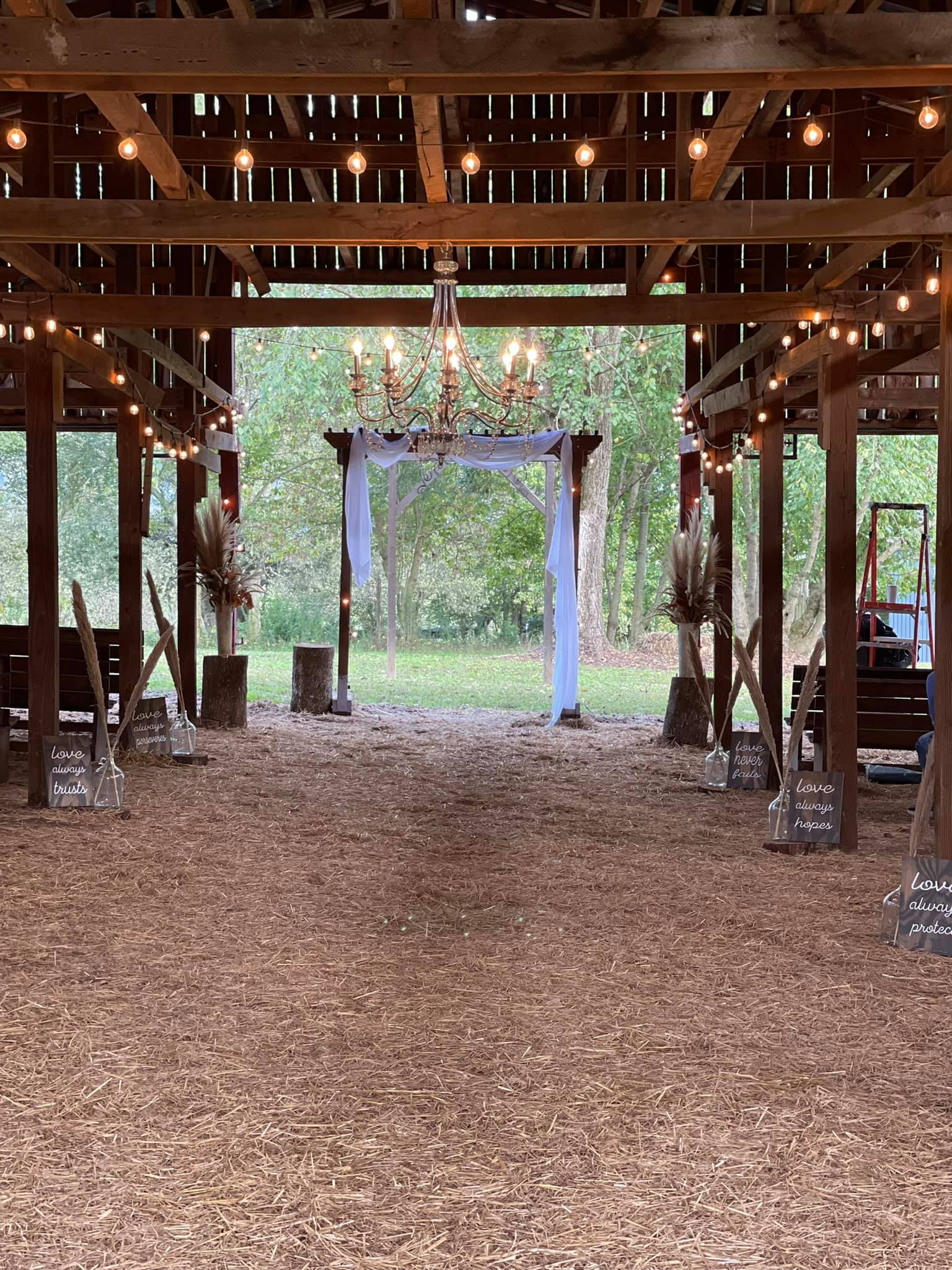 Photo of chandelier and accent lighting in the barn with decorations at the end of each seating row including a hand lettered sign that describes Love. the alter area is trimmed with white bunting. The back of the barn is open to the outside greenspace